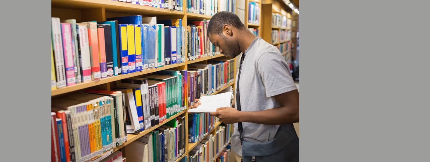 SECONDARY Hero 1440X545px Library AW A young man in a grey t-shirt reading a book in a library aisle